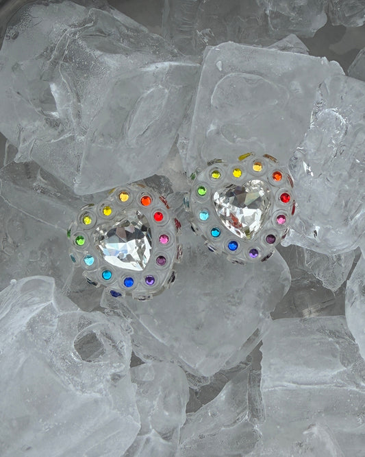 A pair of transparent heart earrings with a central clear heart-shaped crystal and a halo of small multicolored round rhinestones, resting on a bed of ice cubes.