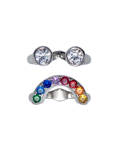 Silver ring with two zircon gemstones and another arch shaped ring with rainbow-colored gemstones that together make a sad looking smiley face