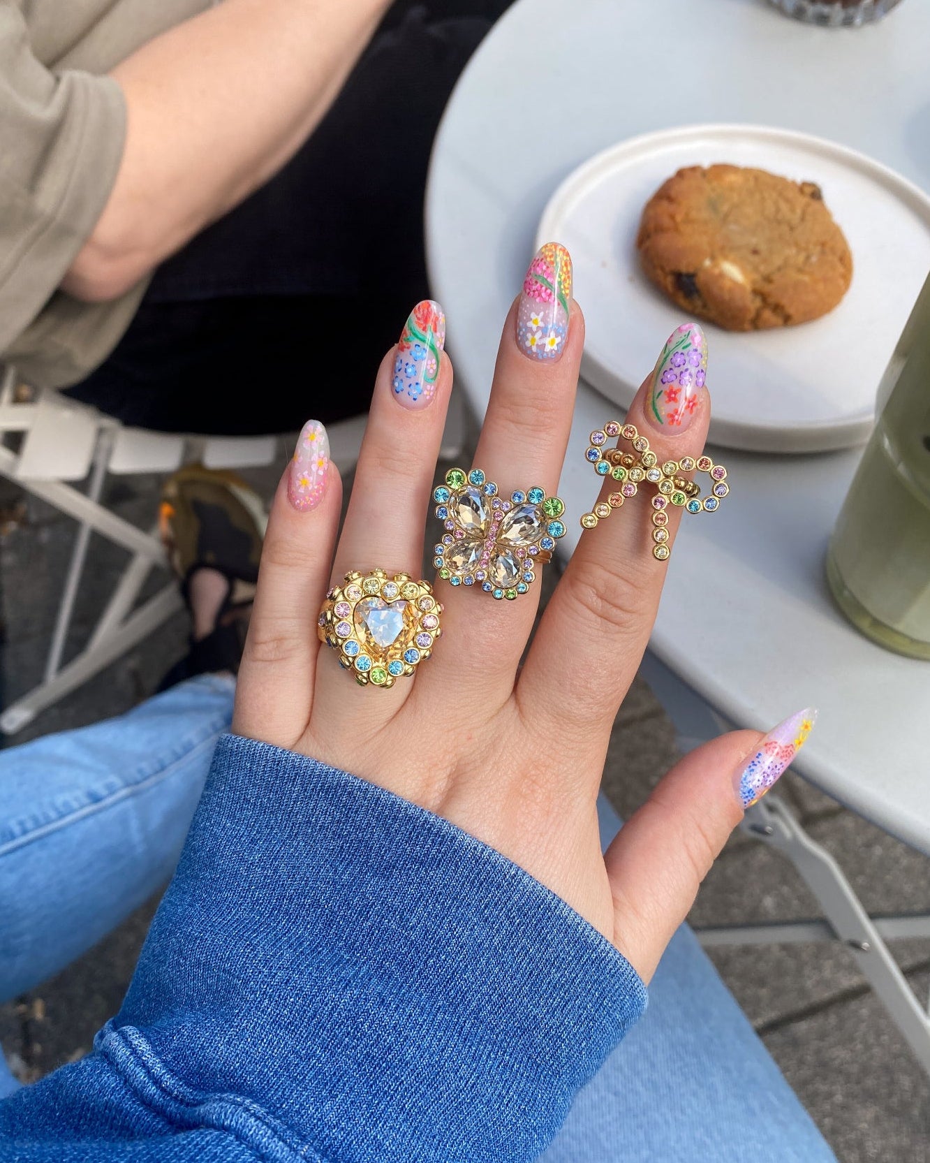 Hand with colorful nail polish and multiple rings, sitting outdoors with a cookie and drink in the background.